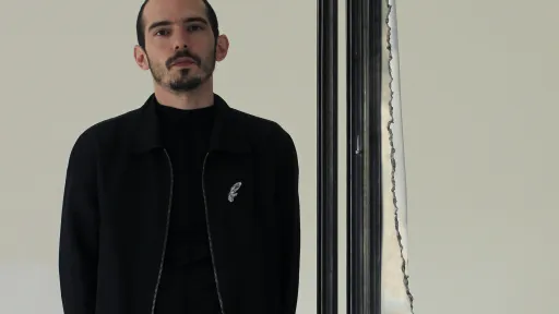 Portrait von Dimitris Tampakis. Der Künstler trägt schwarze Kleidung und blickt durch eine Metallkonstruktion. Im Hintergrund ist eine weisse Wand.