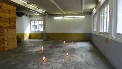 Ein Ausstellungsraum mit 6 brennenden kleinen Objekten auf dem Boden. Die Flammen sind nicht besonders hoch.