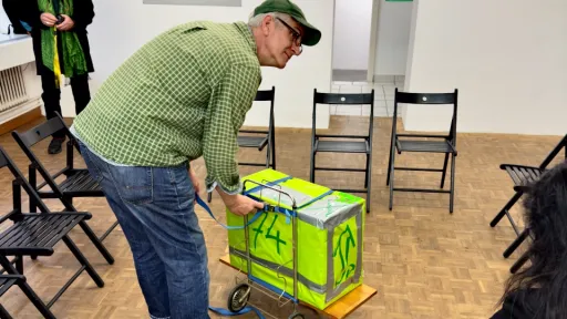 Eine männlich gelesene Person bückt sich zu einer leuchtend grünen Kiste. Um in herum stehen schwarze Stühle.