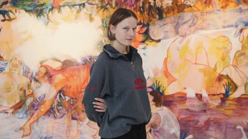 Portrait von Jeanne Jacob. Die Künstlerin steht vor einem farbigen Gemälde und trägt einen grauen Pullover. 