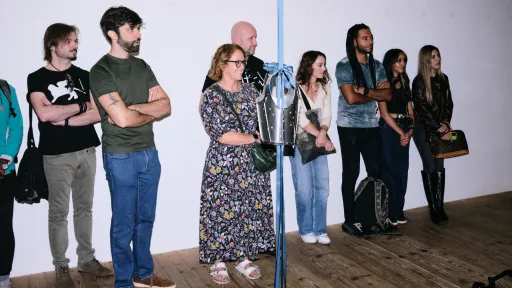 Eine Gruppe steht im Ausstellungsraum. Von der Decke hängt eine Ritterrüstung an einem blauen Satinband