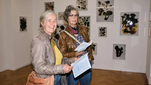 Ausstellungsbesucherinnen blicken lachend in die Kamera. Im Hintergrund hängen Fotografien von Pilzen